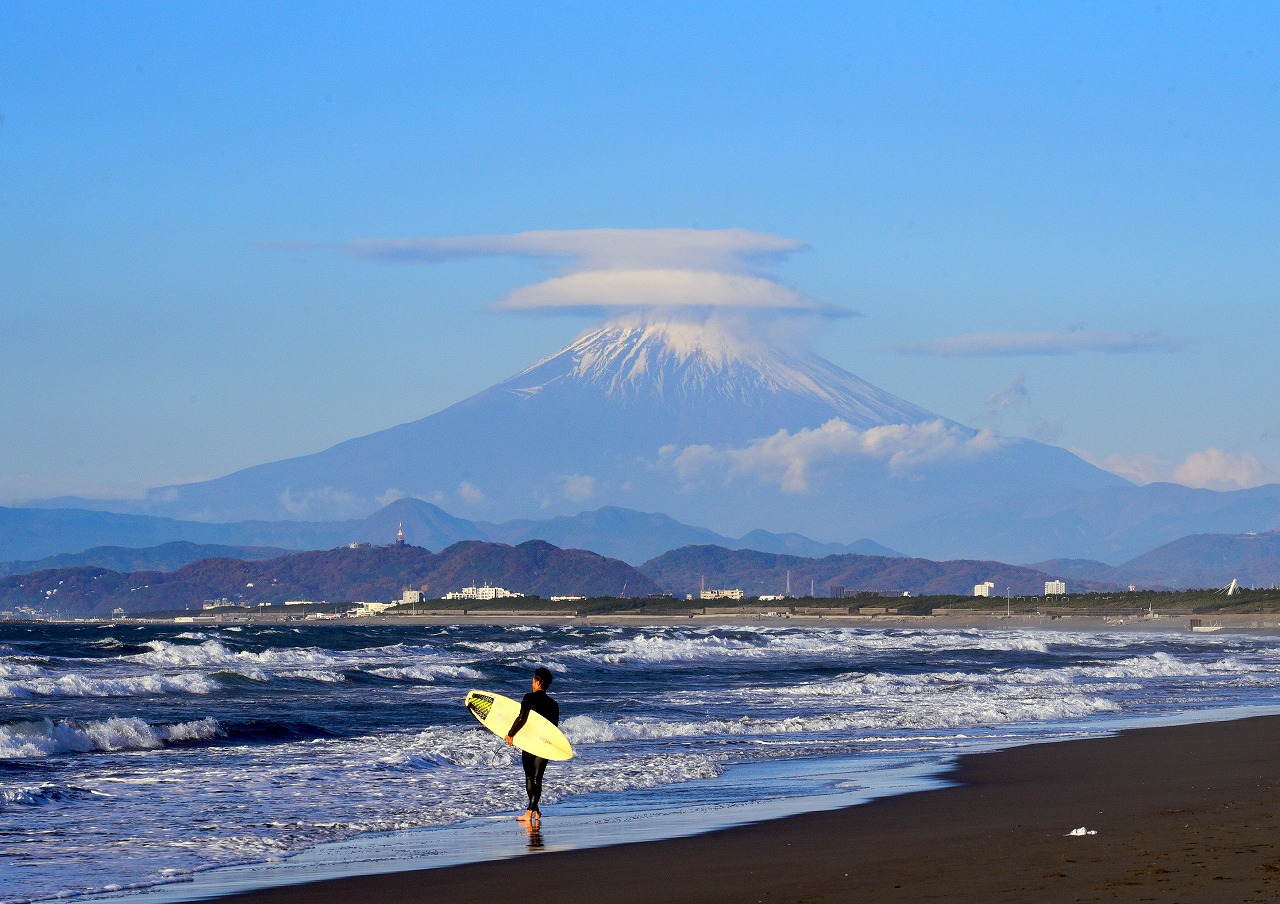 海岸沿いにサーファーが歩いており、背景には笠雲を被った富士山が見える